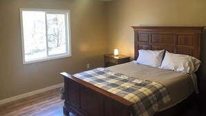 A bedroom with a wooden bed, a nightstand, a window with a view of trees, and a ceiling light. - The Getaway Cabin W/ Indoor hot tub and sauna.
 (Alger)
