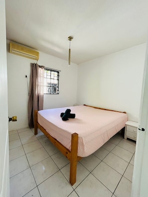 A small, clean room with a single bed, a window with curtains, an air conditioning unit, and a small bedside table.