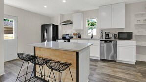 A modern kitchen with white cabinets, a stainless steel refrigerator, a dishwasher, and a coffee maker. There are three black bar stools at the island. - Beautiful 3500 SqFt Villa in Fountain Sq (Indianapolis)