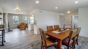 Un comedor amplio con una mesa de madera y sillas de cuero, una cocina al fondo y vistas al exterior a través de ventanas grandes.
