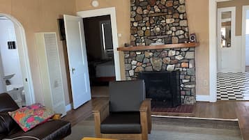 Una sala de estar con chimenea de piedra, ventilador de techo, mesa de centro de madera, sofá de cuero y una puerta que da a otra habitación.