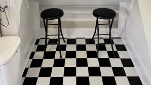 Una cocina con suelo a cuadros blanco y negro, dos taburetes, una botella de vino y un cuadro decorativo en la pared.