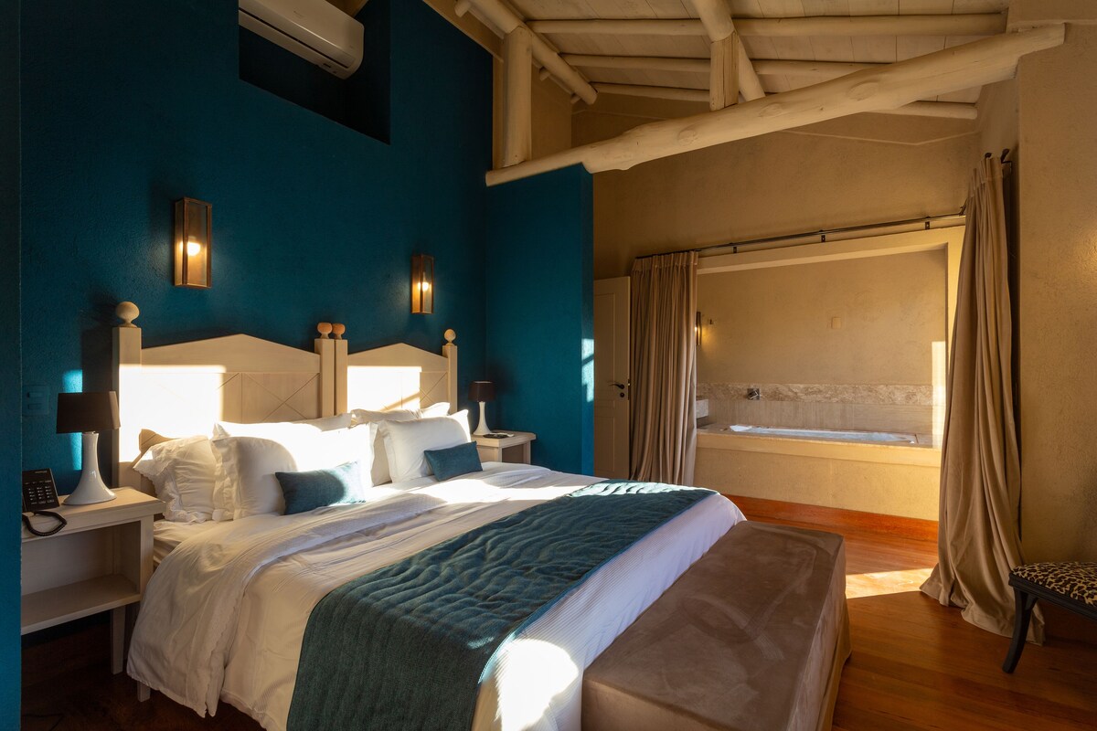 Une chambre à coucher avec un grand lit, deux tables de chevet avec des lampes, une fenêtre avec des rideaux et un plafond en bois.