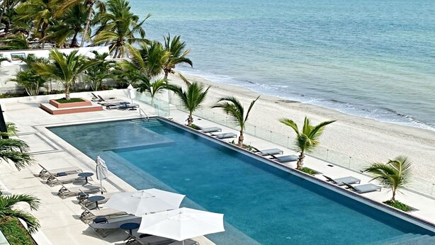 Une piscine rectangulaire située à côté d’une plage, bordée de palmiers et dotée de chaises longues.