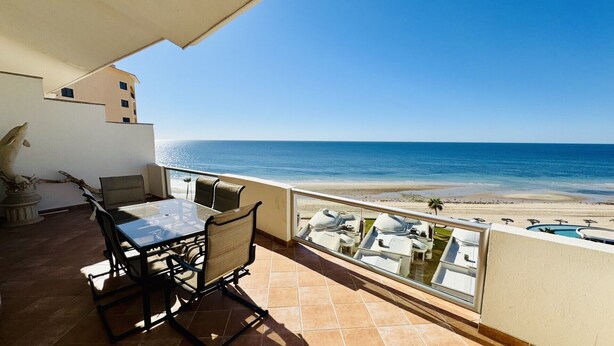 En balkon med et bord og stole, der giver udsigt over en strand og en swimmingpool.