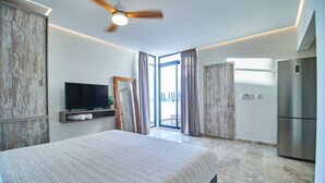A modern hotel room with a large bed, a flat-screen TV, a ceiling fan, and a view through a glass door.