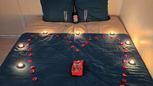Un lit décoré de pétales de rose et de bougies disposés en forme de cœur, une bouteille de champagne et deux verres posés sur la tête de lit.