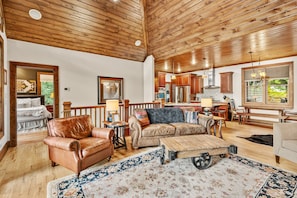 A spacious living area with a high wooden ceiling, a large sofa, a coffee table, and a view of the kitchen.