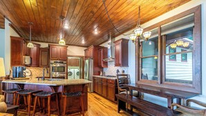 Una cocina con armarios de madera, una isla grande con taburetes de bar, una mesa de comedor con bancos y vistas amplias por grandes ventanales.