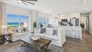 A modern living room with a large window, a ceiling fan, and a kitchen area with stainless steel appliances.