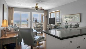 A spacious living room with a large window offering a view of the beach, a ceiling fan, a dining area with a TV, and a kitchen island. - Direct Gulf Views, Private Pool, Sleeps 4 (Fort Walton Beach)