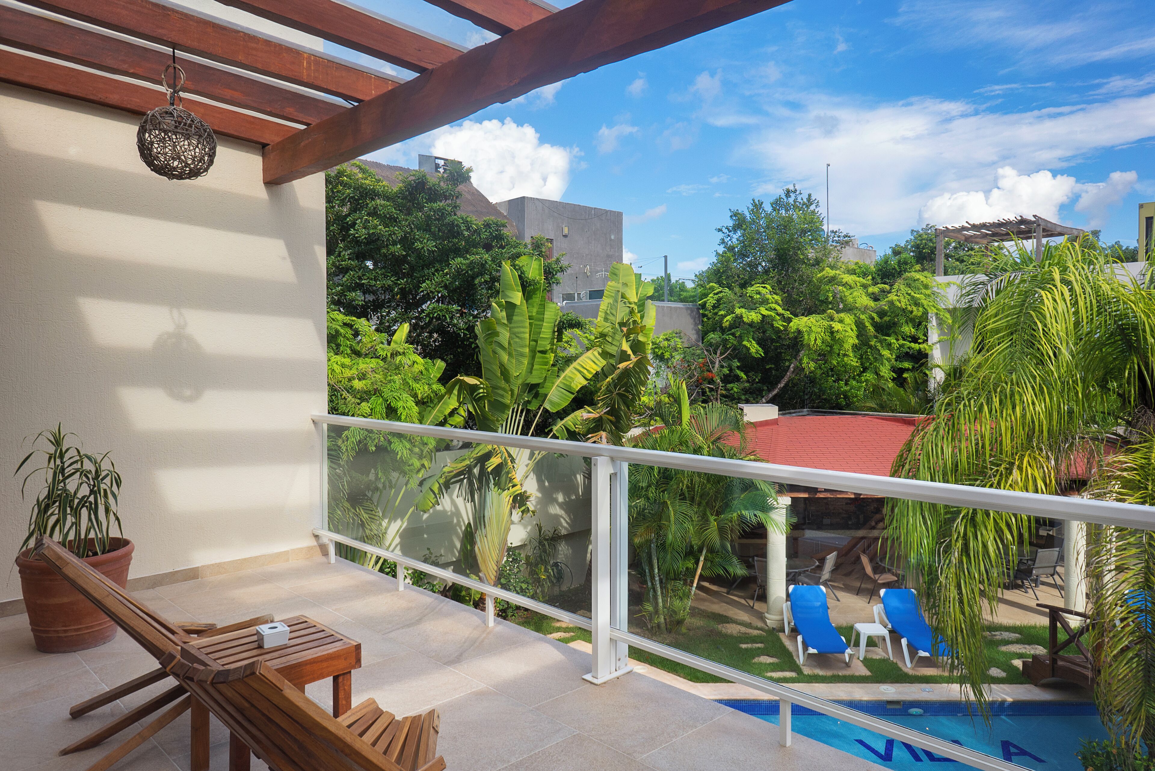 A balcony with a wooden table and chairs, a potted plant, and a view of a swimming pool and greenery.