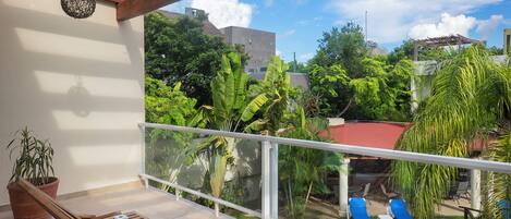 A balcony with a wooden table and chairs, a potted plant, and a view of a swimming pool and greenery.