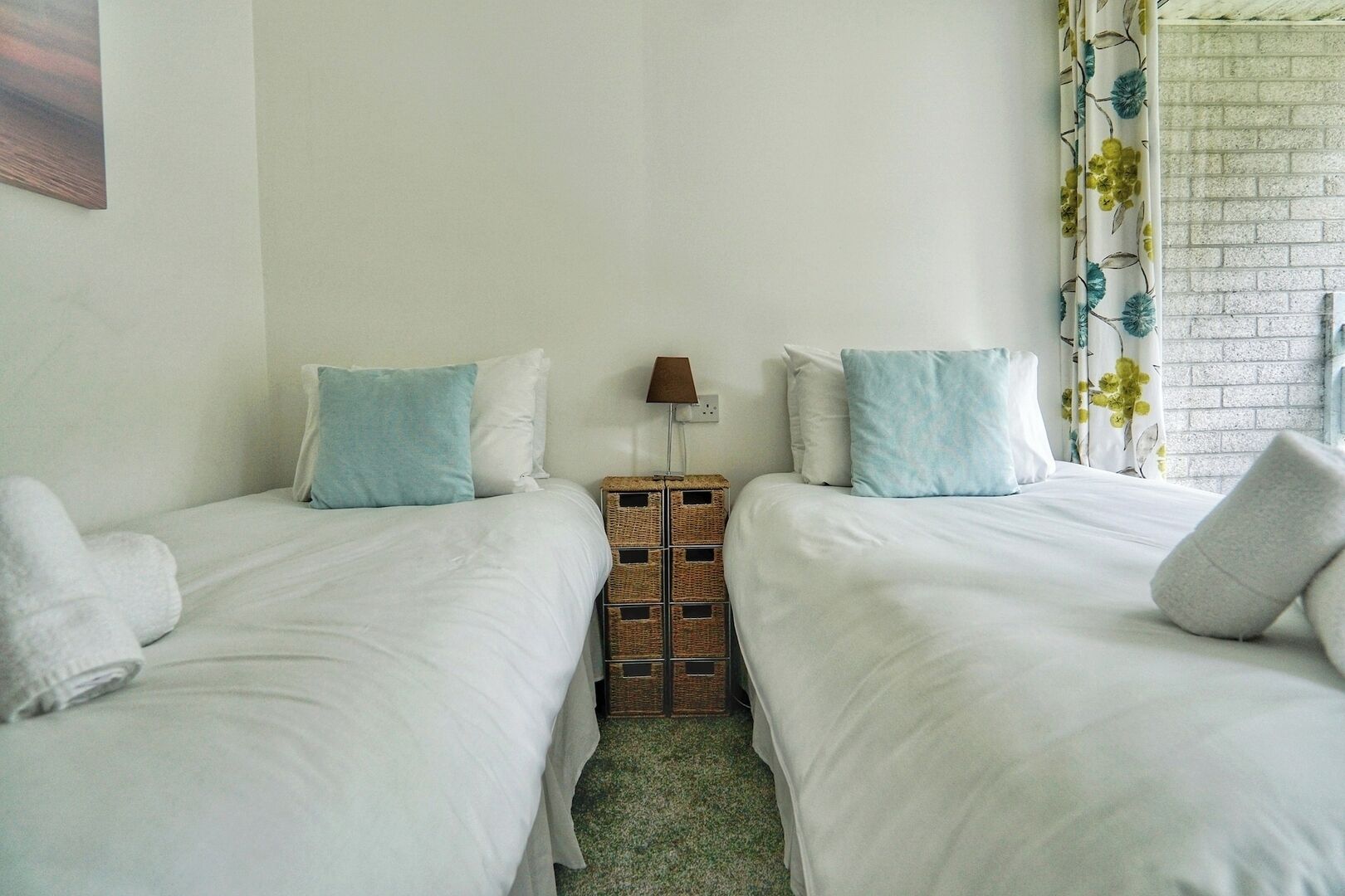 Een hotelkamer met twee bedden, elk voorzien van witte lakens en blauwe kussens. Tussen de bedden staat een kleine tafel met een lamp. De kamer heeft een bakstenen muur en een raam met bloemen gordijnen.
