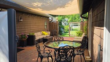 Une terrasse extérieure avec une table ronde et des chaises, un canapé avec des coussins et un sol en briques.