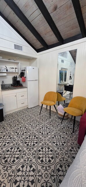 A compact kitchen with a refrigerator, coffee maker, and a sink. There are two chairs and a small table with a mirror above. The floor has a patterned tile design. - The Elderberry Studio (Wanchese)