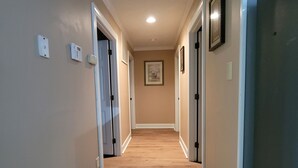 Corredor com paredes bege, detalhes em branco e piso de madeira. Há duas portas à esquerda, uma com interruptor de luz e outra com detector de fumaça. Um quadro emoldurado está pendurado na parede.