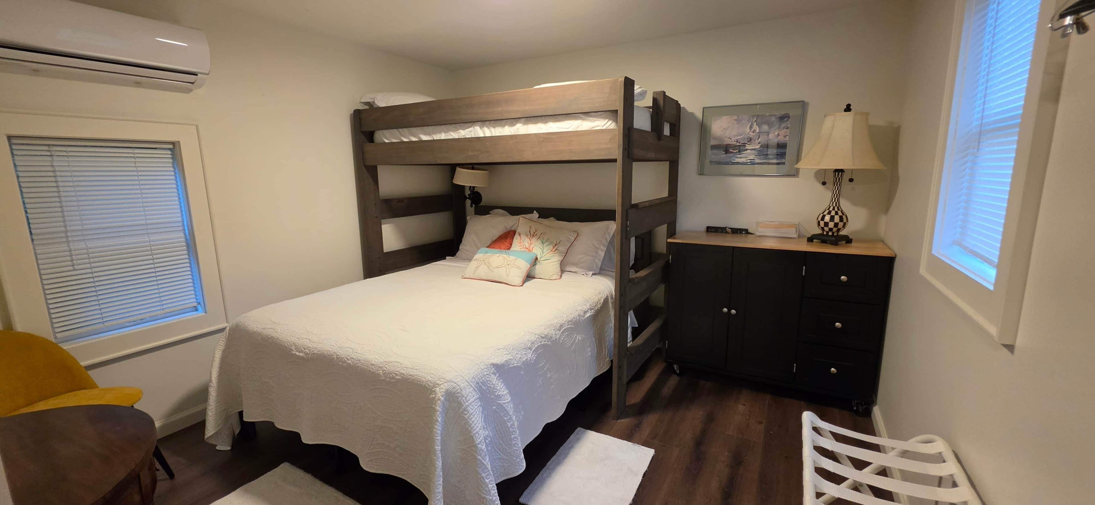 Ein Etagenbett mit weißer Bettdecke, eine Nachttischlampe, ein kleiner Schrank und ein Fenster mit Jalousien.