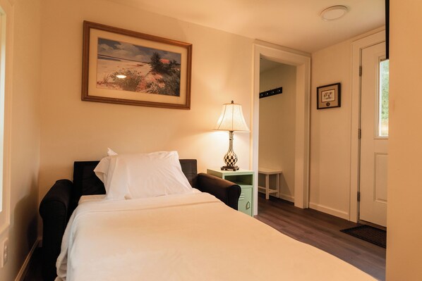 A bedroom with a bed, a nightstand, a lamp, a framed picture on the wall, and a door leading to another room.