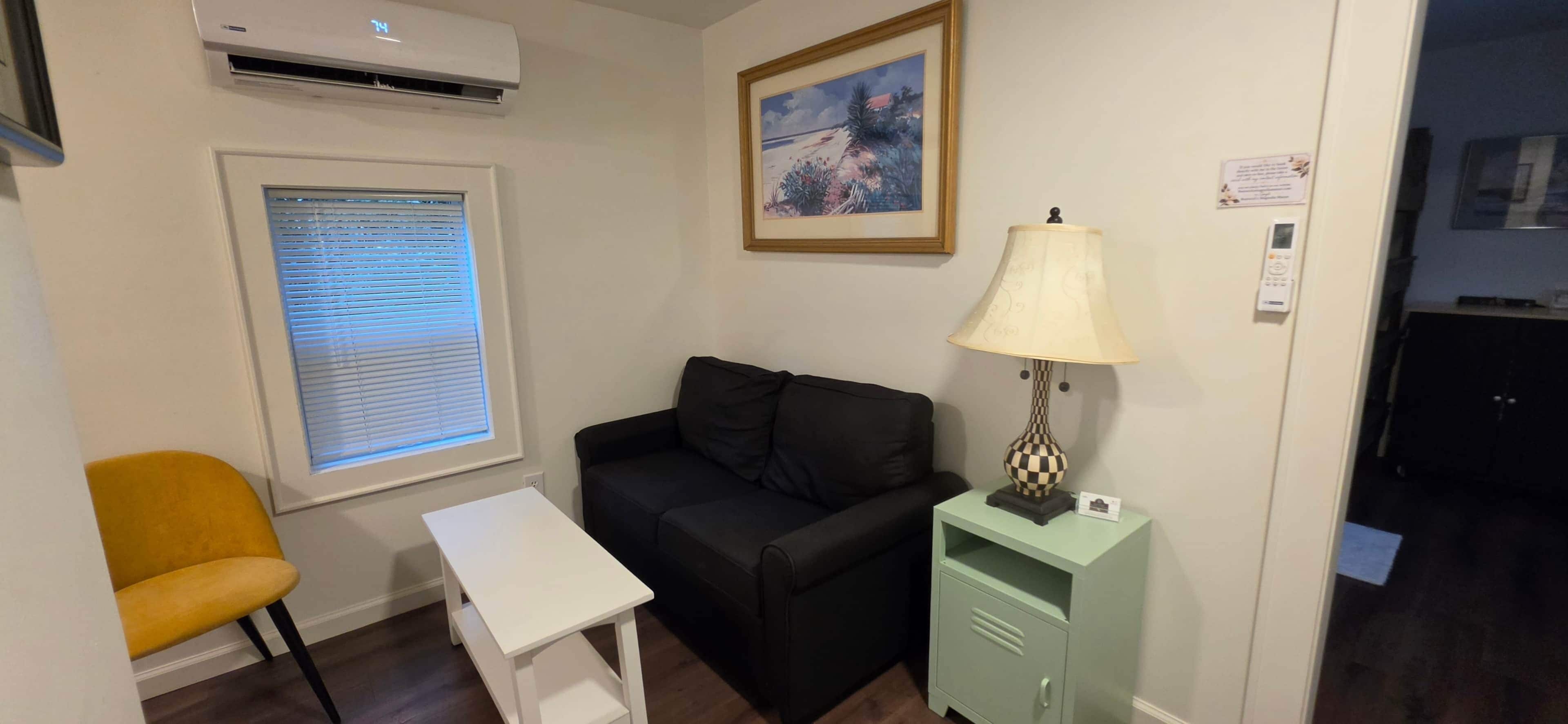 Ein kleiner Raum mit einem schwarzen Sofa, einem gelben Sessel, einem weißen Tisch, einer Lampe und einem gerahmten Bild an der Wand.
