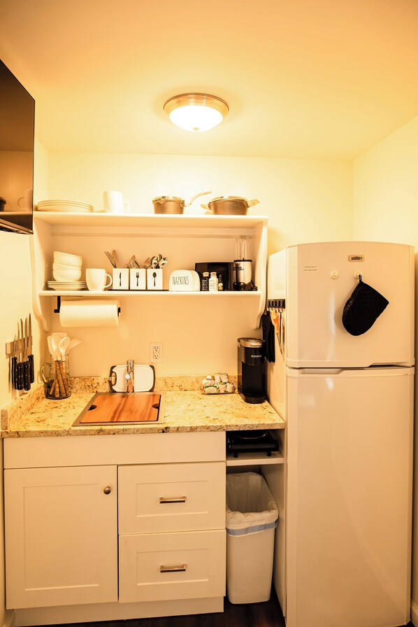 Una cocina con armarios blancos, una encimera con tabla de cortar, un fregadero, un cubo de basura y un refrigerador.