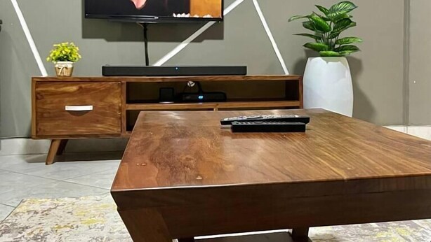 Ein modernes Wohnzimmer mit Couchtisch aus Holz, einem flachbildfernseher an der Wand, einer kleinen Pflanze und einer Wand mit geometrischen Mustern.