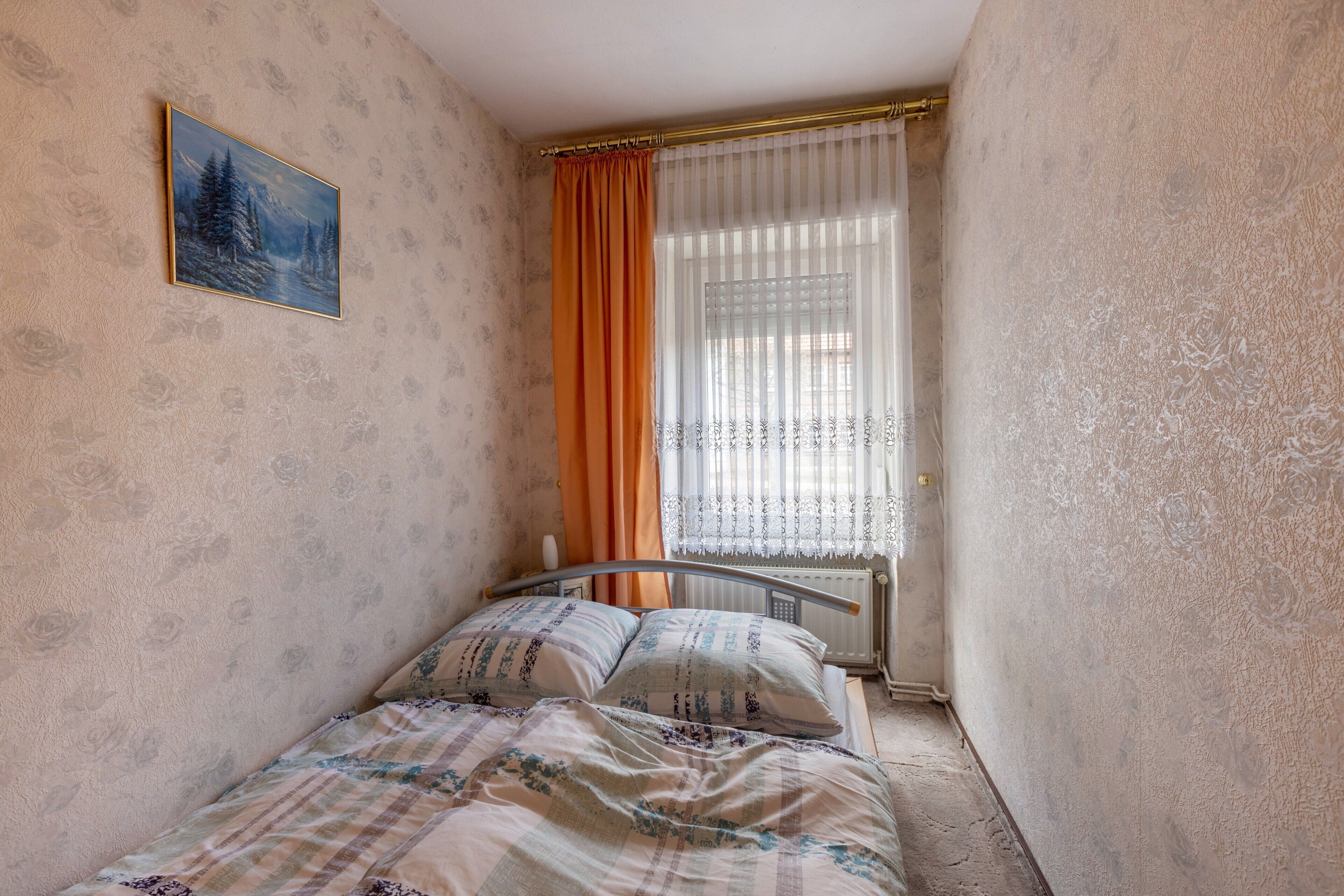 Ein schmaler Raum mit einem Bett, einem Heizkörper und einem Fenster mit Vorhängen.