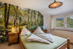 Ein Schlafzimmer mit großem Wandgemälde von einem Wald, einem Bett mit Kissen, einem Nachttisch und einem Fenster mit Blick.