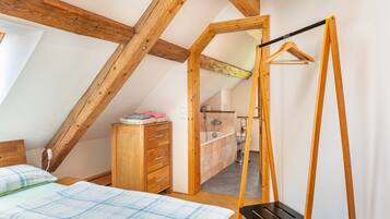 Una camera da letto con soffitto spiovente, travi in legno, un letto con copriletto a quadri, un comò in legno e un bagno visibile attraverso una porta aperta.