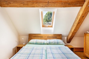 Une petite chambre à coucher accueillante, avec un plafond en pente, un lit simple avec une couette à carreaux, une table de chevet avec une lampe et une fenêtre donnant sur les arbres.