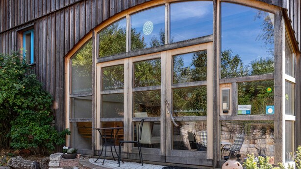 A wooden building with large windows, outdoor seating, and a stone wall. - Holiday Apartment Wegwarte with Mountain View, Shared Terrace and Wi-Fi (Weilheim)