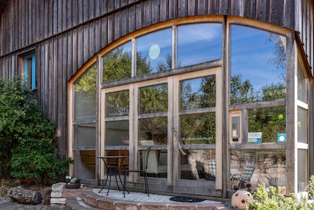 A wooden building with large windows, outdoor seating, and a stone wall.. Holiday Apartment Wegwarte with Mountain View, Shared Terrace and Wi-Fi