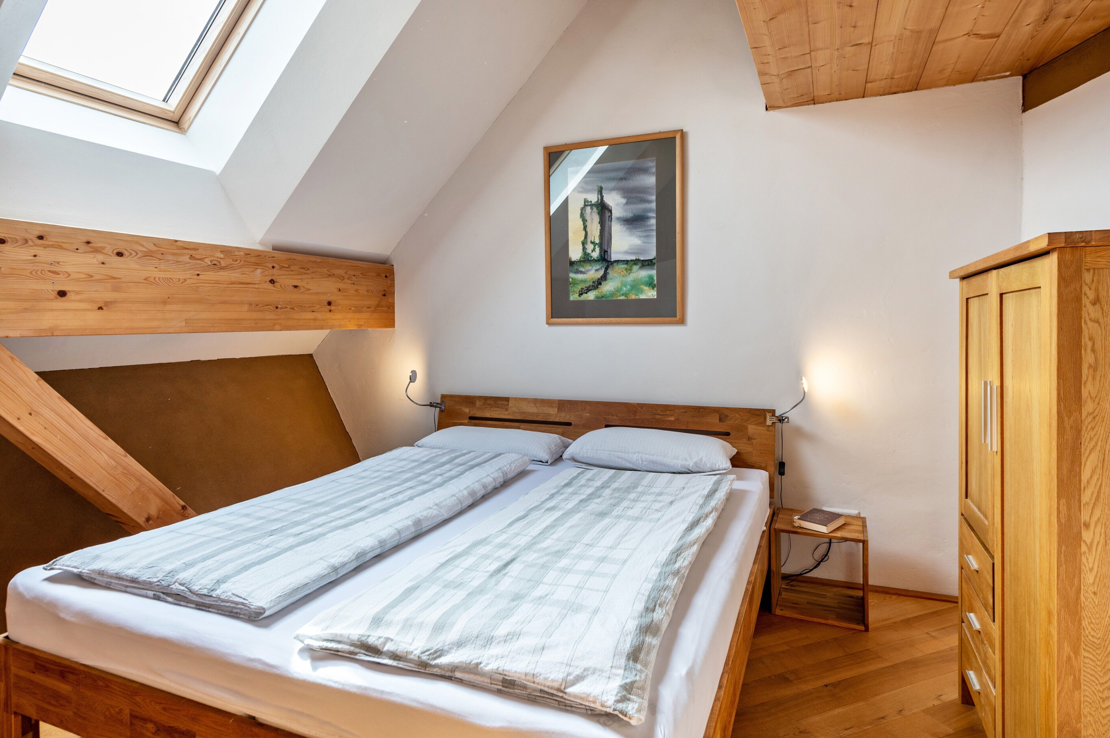 Un dormitorio con techo inclinado, una cama con cabecera, una mesita de noche, un armario de madera y un cuadro en la pared.
