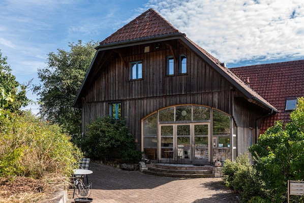 A wooden building with a sloped roof, large windows, and a paved entrance area with outdoor seating.