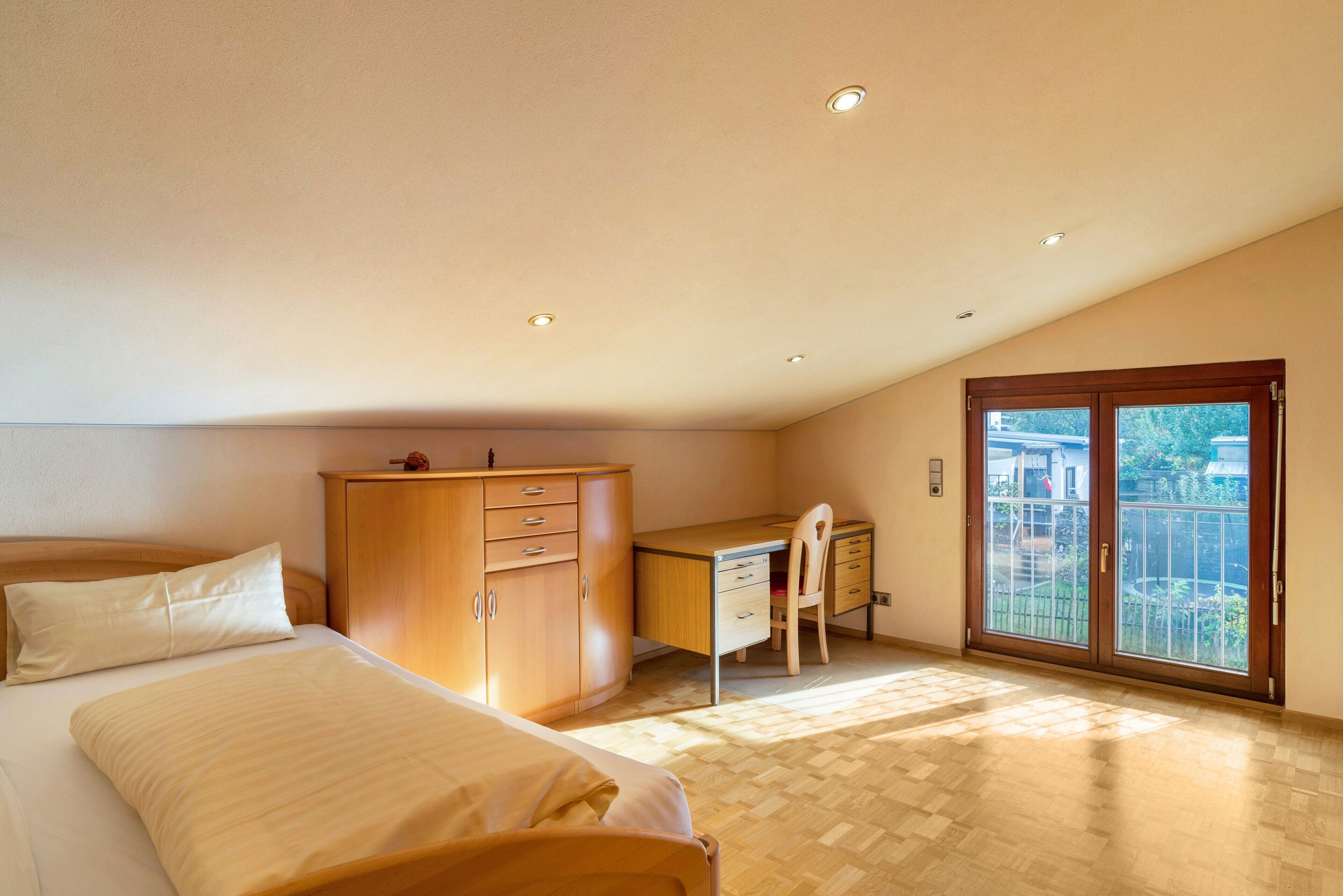 Une chambre à coucher comprenant un lit, un bureau, une chaise, une armoire et une porte-fenêtre coulissante donnant sur un balcon.