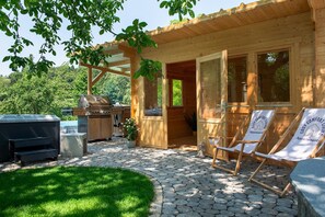 Una cabaña de madera con un patio de piedra, una parrilla y dos sillones de descanso.