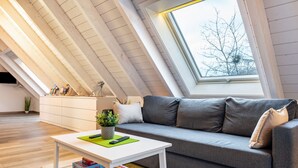 An attic room with a sloped ceiling, a grey sofa, a white coffee table, and a window with a view of bare trees.