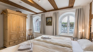 Une chambre avec une grande armoire en bois, deux lits avec du linge de lit blanc, un tableau au mur et de grandes fenêtres avec des rideaux.
