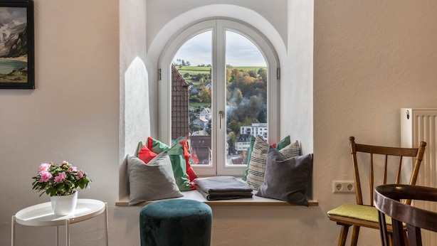 Une pièce dotée d’une grande fenêtre en arc de cercle, d’une petite table sur laquelle est posée une plante en pot, d’un banc rembourré et d’une chaise en bois.