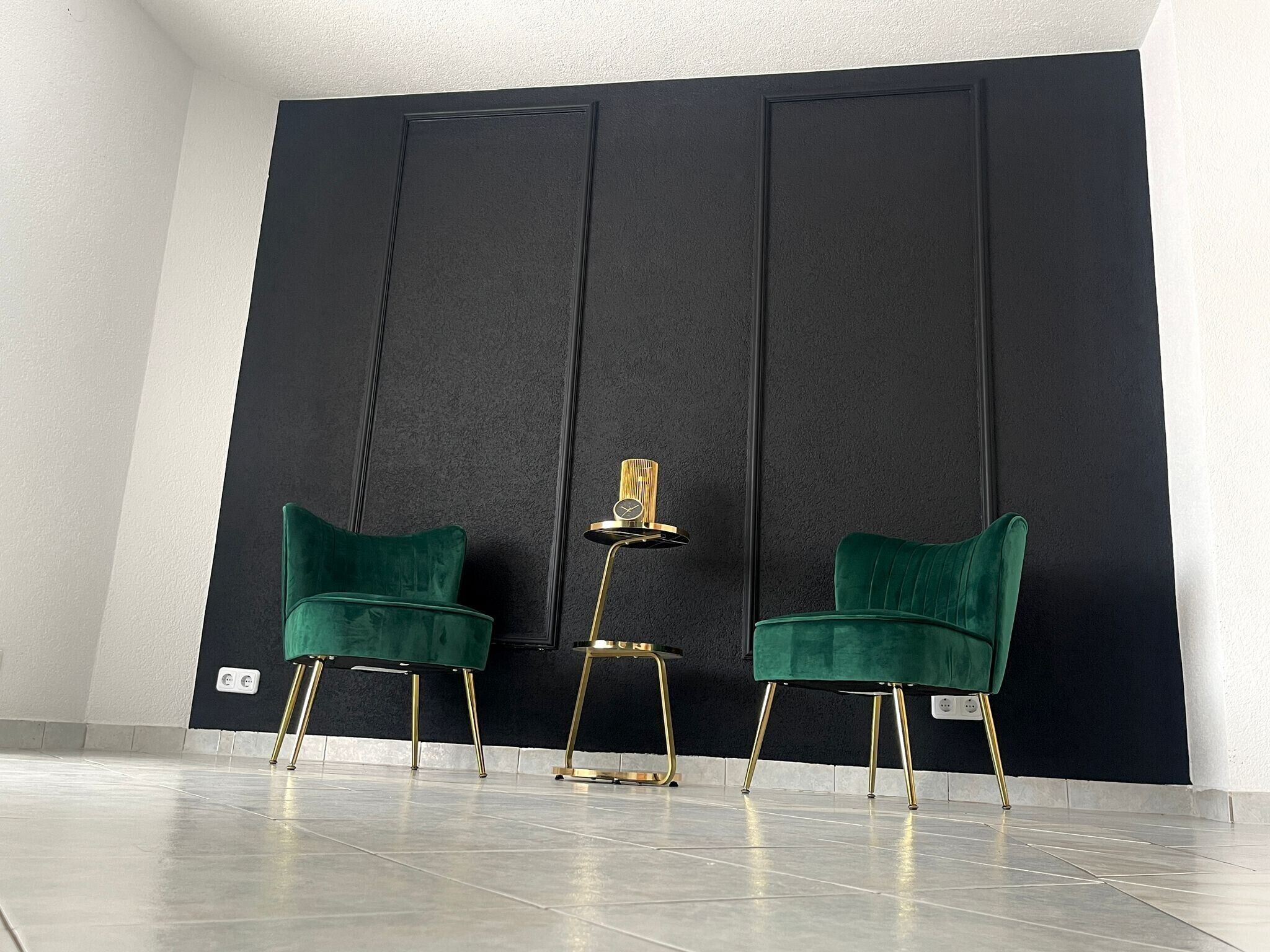 Una habitación con dos sillones de terciopelo verde, una mesita con un reloj y una pared oscura con líneas verticales.
