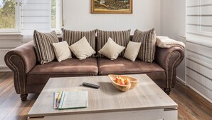 A brown leather sofa with striped pillows, a wooden coffee table with a bowl of fruit, a remote control, and a book. - Apartment 'Ferienwohnung 2, 80qm' with Mountain View, Shared Garden and Wi-Fi (Rosswangen)