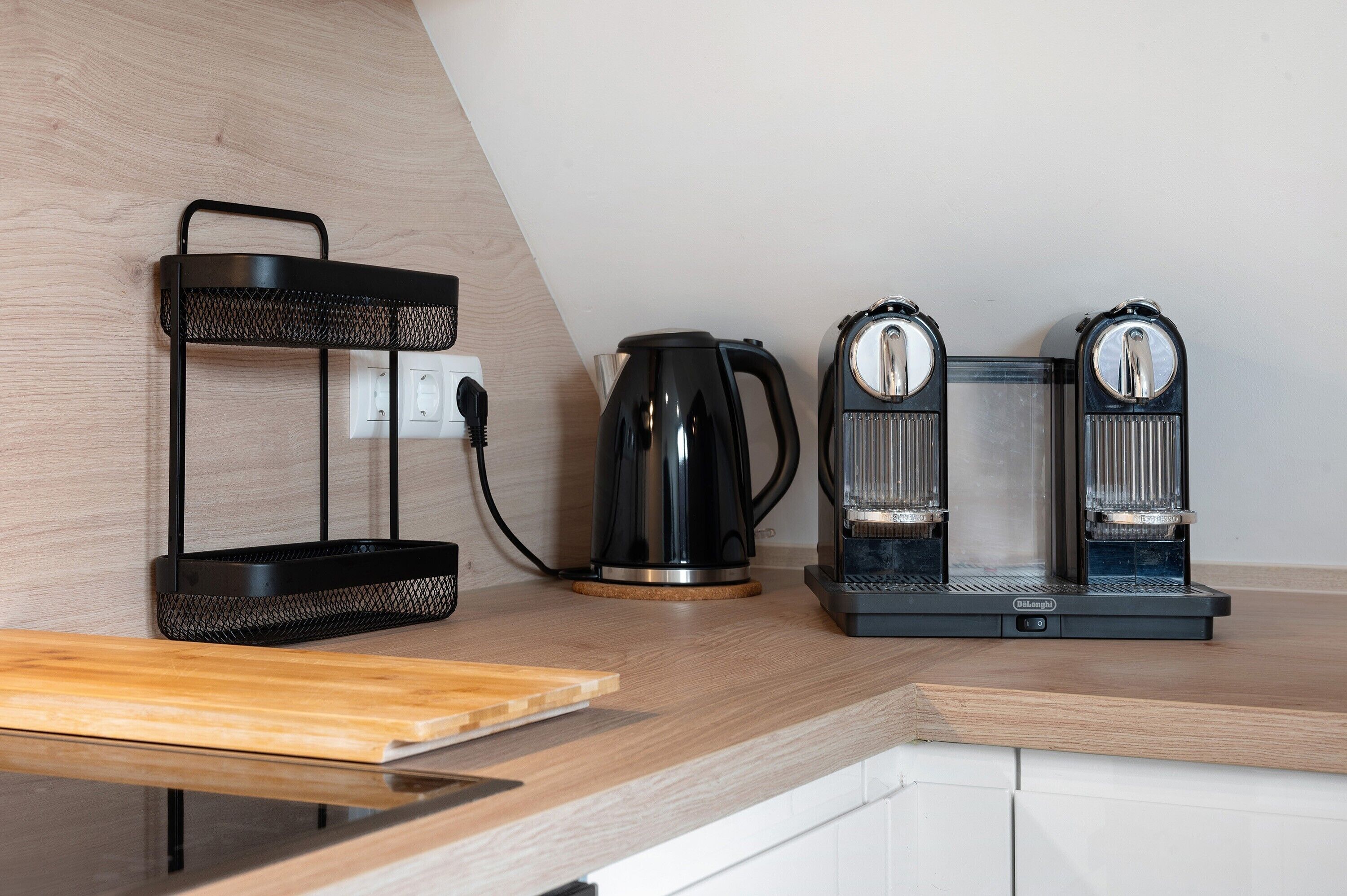 Une cuisine équipée d’une machine à café, d’une bouilloire et d’une étagère en métal noir posée sur un plan de travail en bois.