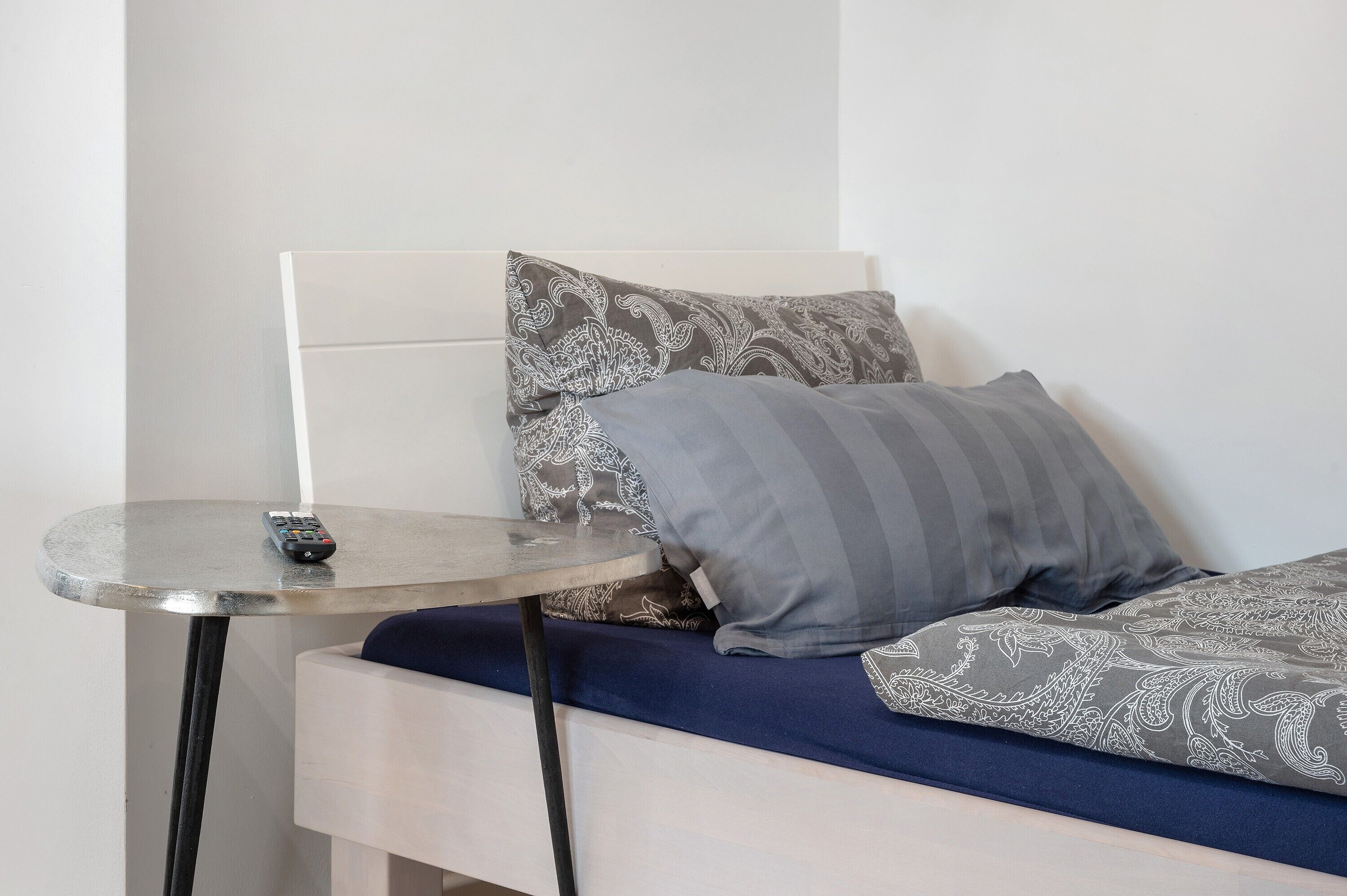 Ein Bett mit gemusterten Betttextilien, eine Fernbedienung auf einem kleinen Tisch daneben und ein weißes Kopfteil.