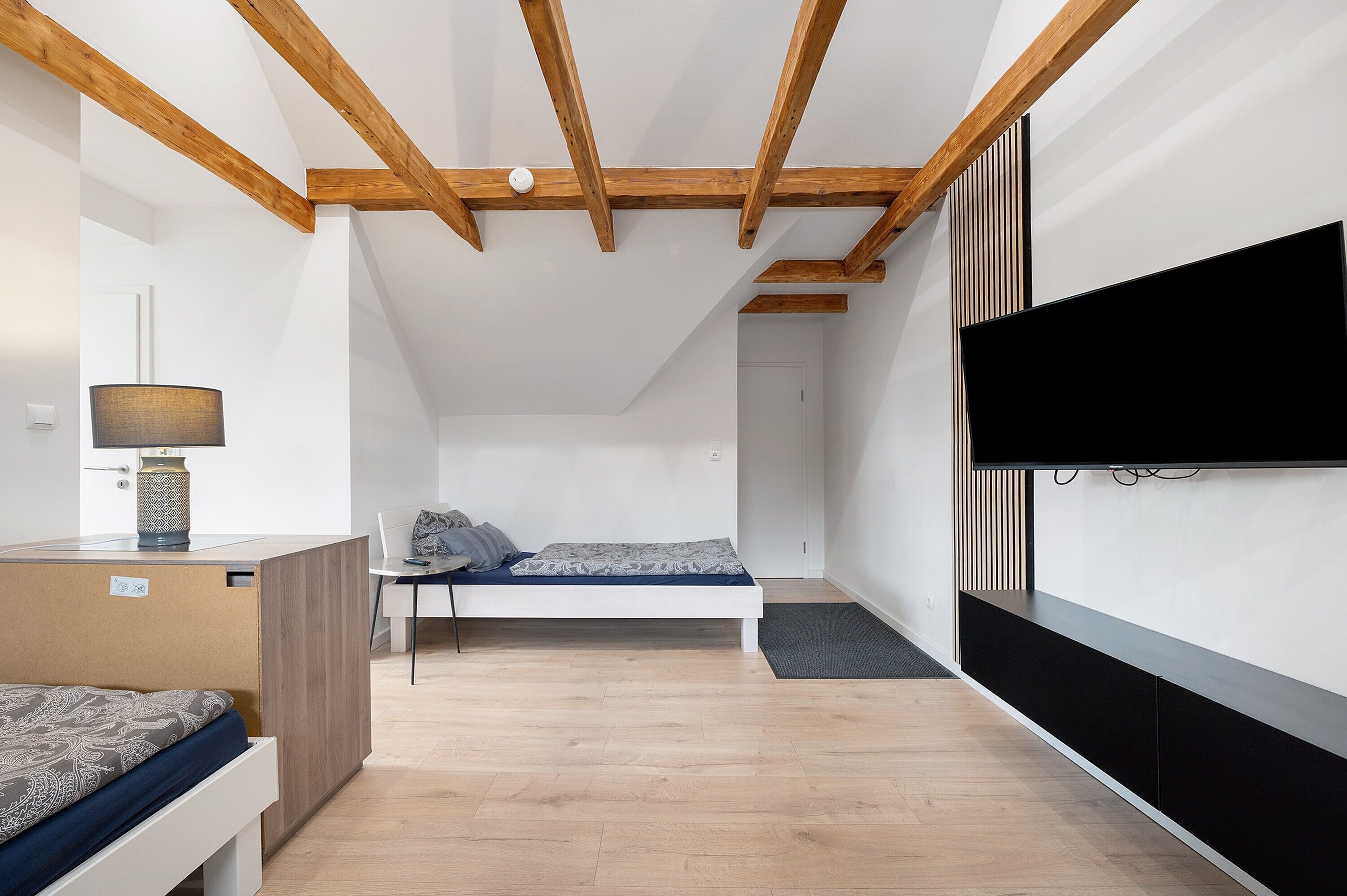 A modern bedroom with a bed, a desk, a lamp, a TV, and a wooden ceiling structure.