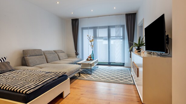 Una sala de estar moderna con un sofá, una cama, un televisor y un amplio ventanal.