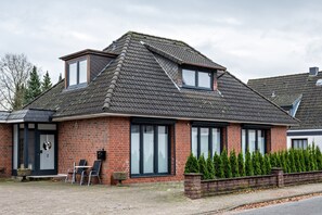 Une maison en briques avec un toit en pente, un petit garage attenant et un porche à l’avant, aménagé avec des sièges pour l’extérieur.