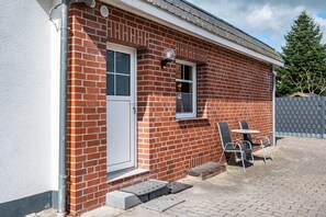 Un bâtiment en briques rouges avec une porte blanche, une fenêtre avec un volet et un petit espace extérieur aménagé avec deux chaises et une table.