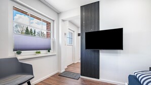 Una habitación moderna con un ventanal grande, un televisor montado, una silla gris y una cama con ropa de cama a rayas.