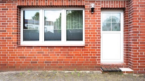Ein Gebäude aus roten Ziegelsteinen mit Fenster- und Türrahmen in Weiß, eine kleine Stufe zum Eingang und ein klarer Himmel, der durch das Fenster sichtbar ist.