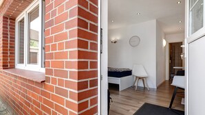 Ein rot backenfassender Außenbau mit Fenster, eine weiße Tür, die in einen Raum mit einem Bett, einem Sessel und einem kleinen Tisch führt.